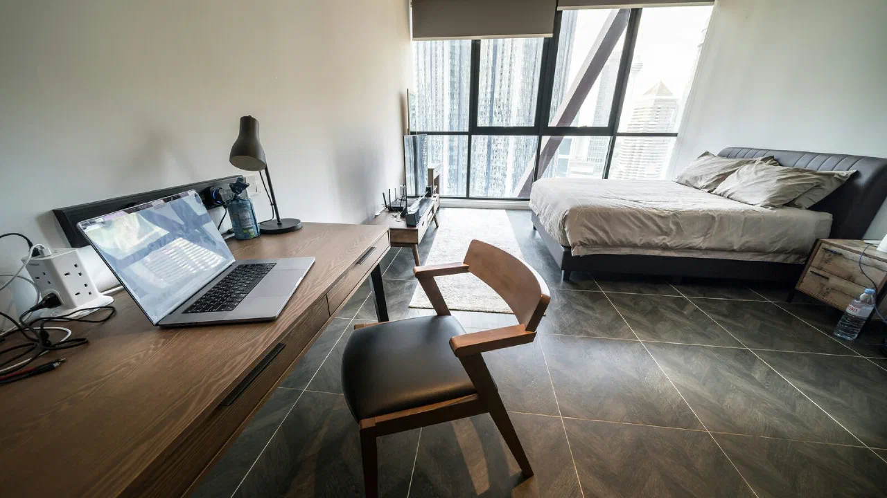 A wooden desk with a laptop and a lamp beside a tall window overlooking a city